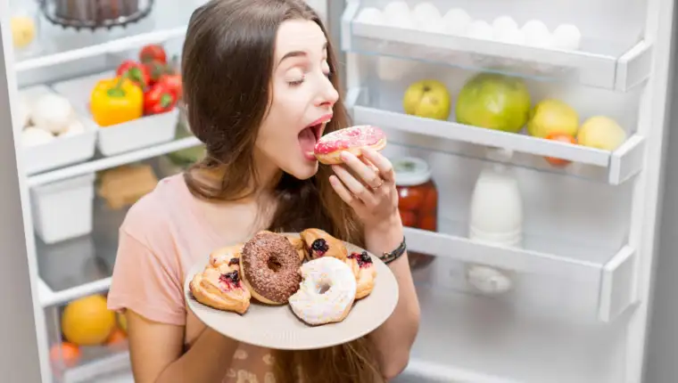 Young woman in the sleepwear eating sweet donuts near the refrigerator