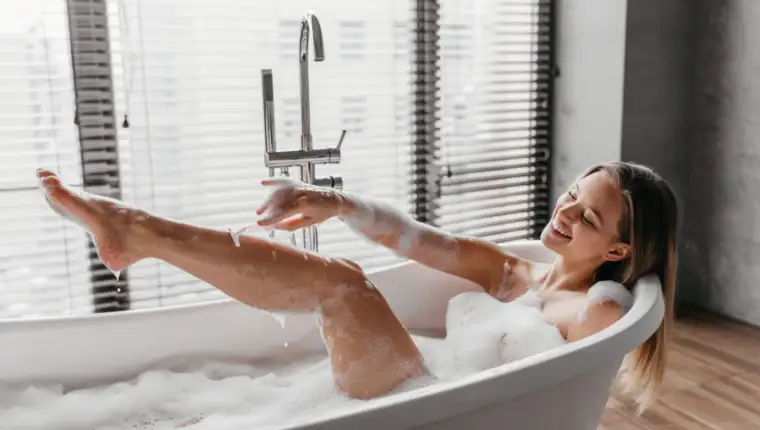 Personal care at home. Happy lady shaving legs with razor, lying in foamy bubble bath, doing skin care procedure