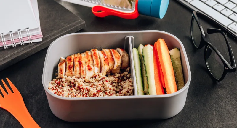 Healthy meal prep containers with quinoa, chicken and and sticks of cucumber and carrots on black wooden background. Healthy lunch at work. Take away concept.
