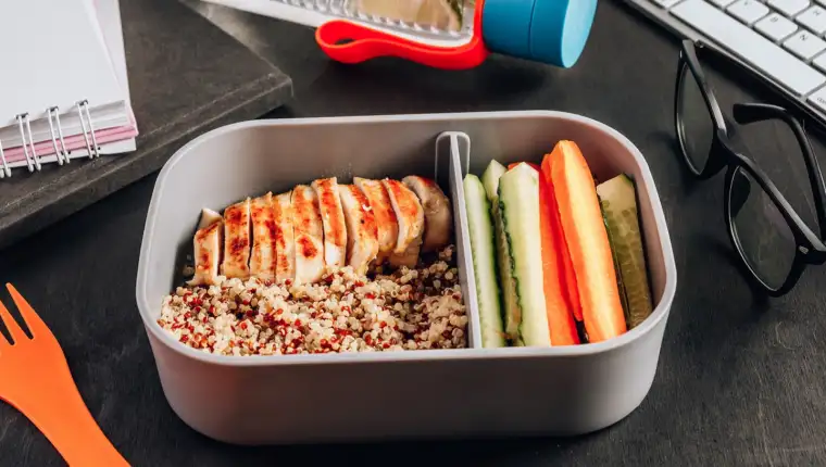 Healthy meal prep containers with quinoa, chicken and and sticks of cucumber and carrots on black wooden background. Healthy lunch at work. Take away concept.