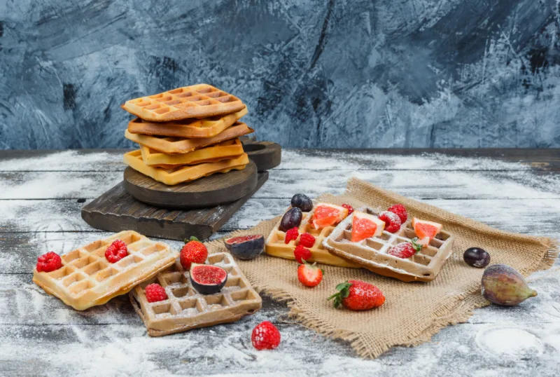 Waffles on wooden board with strawberry,plum,piece of sack on wood with sprinkled flour background, high angle view