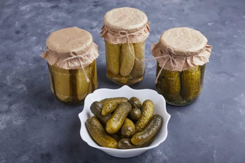 Pickled cucumbers in bowl and glass jars on blue background