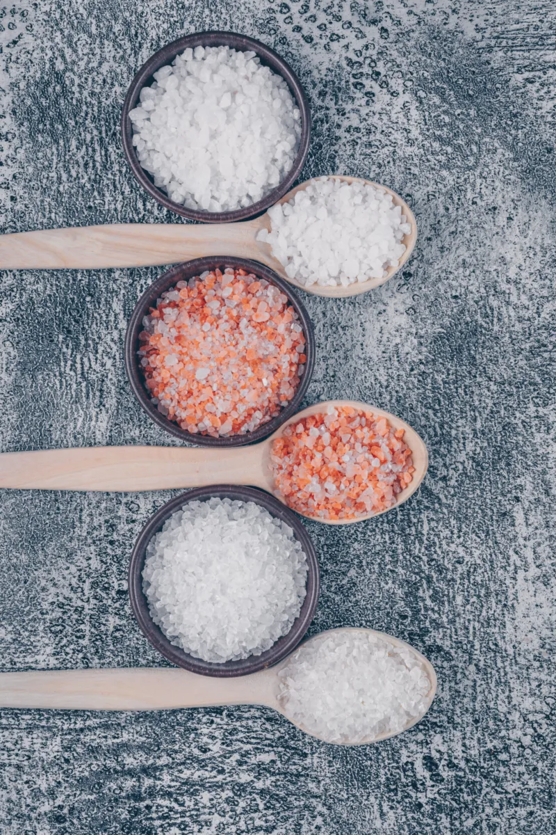 Top view sea salt in bowls and wooden spoons with himalayan salt on gray textured background. vertical