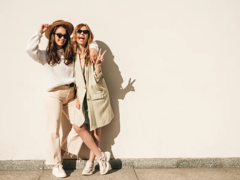 Two young beautiful smiling hipster female in trendy white sweater and coat. Sexy carefree women posing on the street background in hats. Positive models having fun outdoors. Hugging