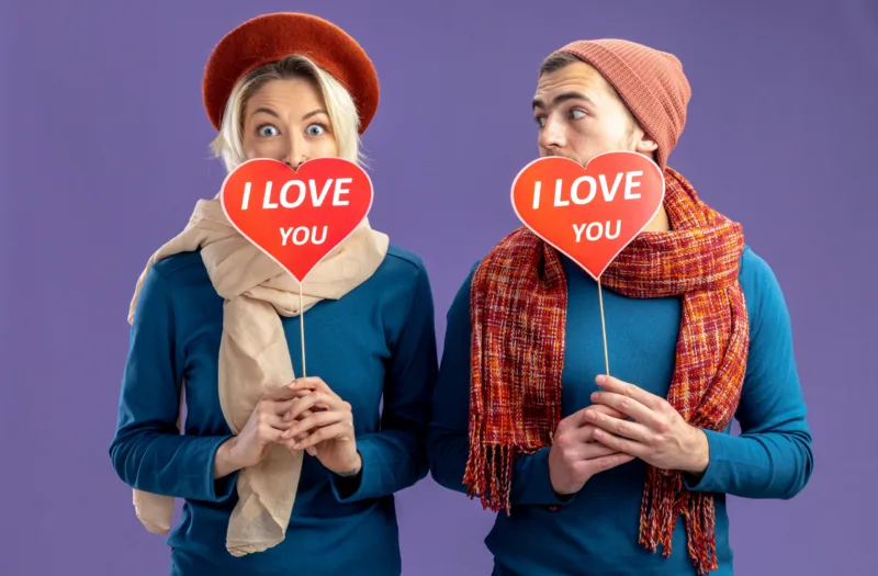 young couple wearing hat with scarf on valentine's day covered face with red hearts on a stick with i love you text pleased guy looking at girl isolated on blue background