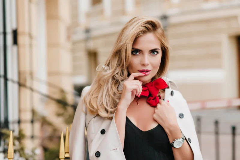 Blue-eyed female model in trendy red scarf posing with pleasure in new outfit standing beside building. Gorgeous fair-haired woman with sparkle make-up touching lip with fingers with surprised smile..