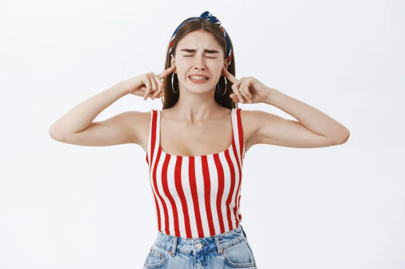 Stylish girlfriend irritated with pissing noise of horrible music being displeased closing eyes and clenchign teeth from dislike closing ears with index fingers standing annoyed over gray background