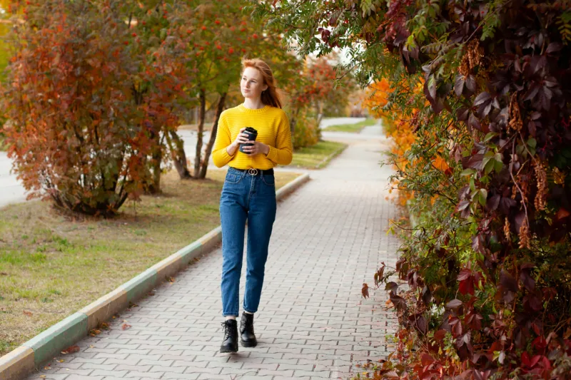 Autumn. A beautiful young red-haired girl with freckles walks down the street with coffee in her hands. Color.