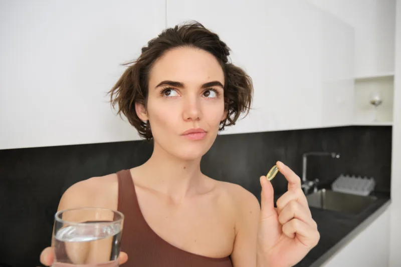 Close up portrait of sportswoman in activewear, holding omega-3, vitamin c, d and b in her hand, taking dietary supplement with drinking glass of water. Healthy lifestyle and sport concept
