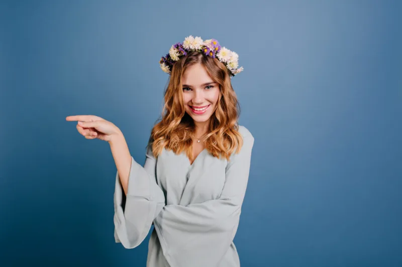 Stunning fair-haired girl in flower wreath posing with playful smile. Studio shot of enthusiastic young woman in romantic attire expressing good emotions..