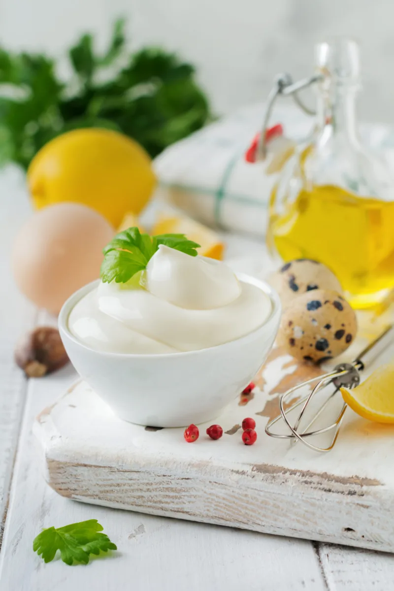 Traditional mayonnaise sauce in white ceramic bowl and ingredients for its preparation on white wooden background. Selective focus