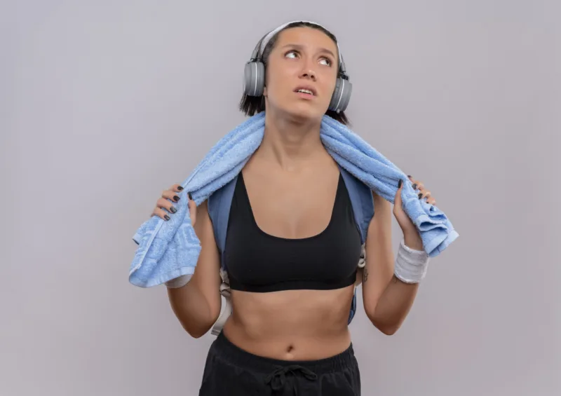 young fitness girl in sportswear with headphones on head and towel on her neck looking aside tired and bored standing over white background