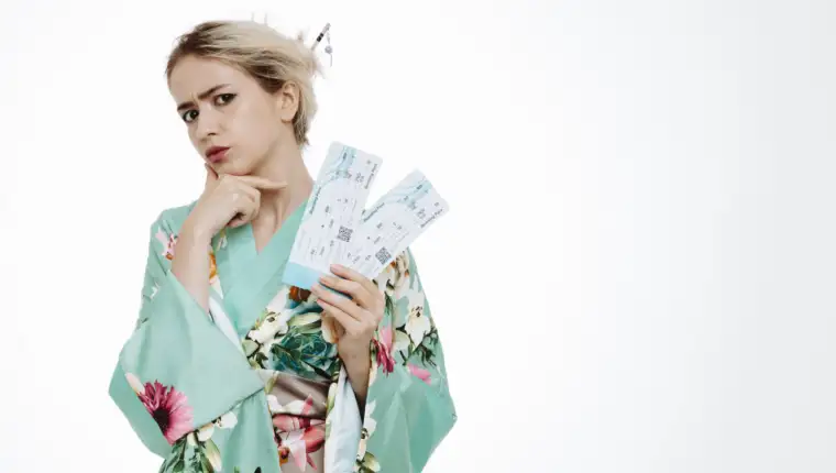 young geisha woman in traditional japanese kimono holding air tickets looking at camera with skeptic expression holding hand on her chin standing over white background
