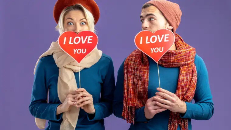 young couple wearing hat with scarf on valentine's day covered face with red hearts on a stick with i love you text pleased guy looking at girl isolated on blue background