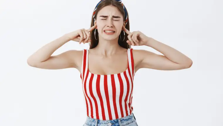 Stylish girlfriend irritated with pissing noise of horrible music being displeased closing eyes and clenchign teeth from dislike closing ears with index fingers standing annoyed over gray background