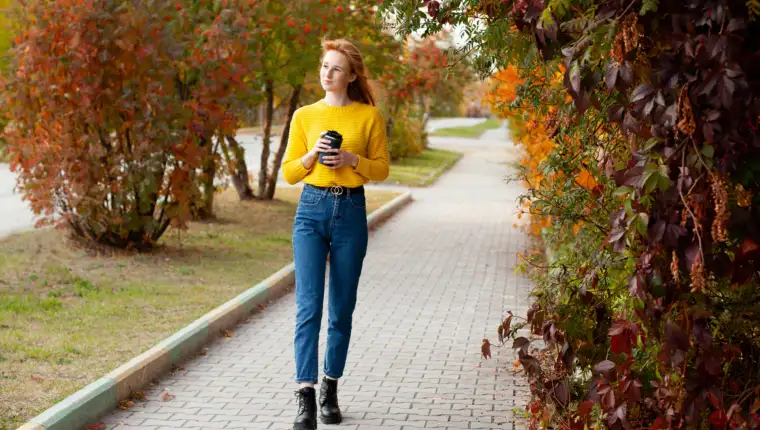 Autumn. A beautiful young red-haired girl with freckles walks down the street with coffee in her hands. Color.