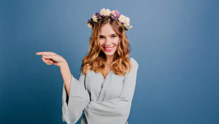Stunning fair-haired girl in flower wreath posing with playful smile. Studio shot of enthusiastic young woman in romantic attire expressing good emotions..