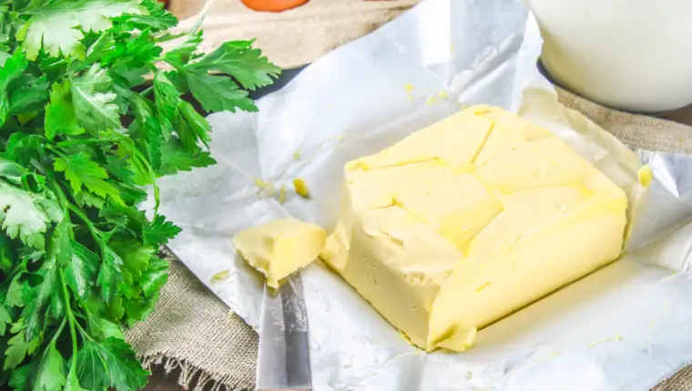 A bar of butter is cut into pieces on a wooden board with a knife, surrounded by milk, eggs and parsley on a brown table. Ingredients for cooking.