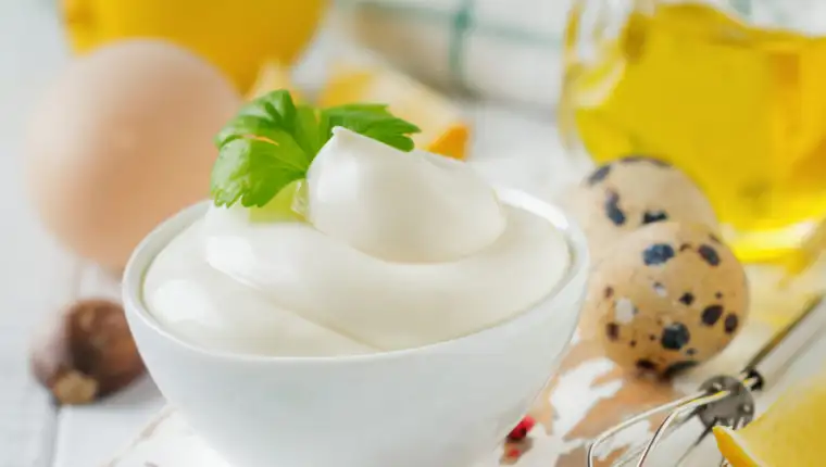 Traditional mayonnaise sauce in white ceramic bowl and ingredients for its preparation on white wooden background. Selective focus