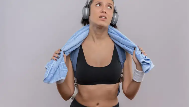 young fitness girl in sportswear with headphones on head and towel on her neck looking aside tired and bored standing over white background