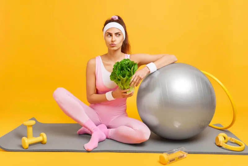 Healthy slim woman keeps to diet has regular fitness training for keeping fit holds green vegetable dressed in sportsclothes sits on mat with headphones dumbbells swiss ball hula hoop in gym