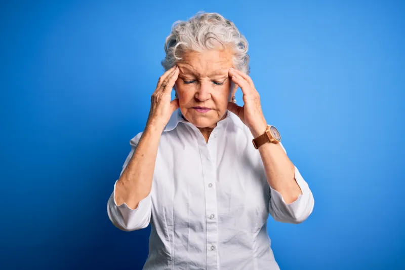 Senior beautiful woman wearing elegant shirt standing over isolated blue background with hand on head for pain in head because stress. Suffering migraine.