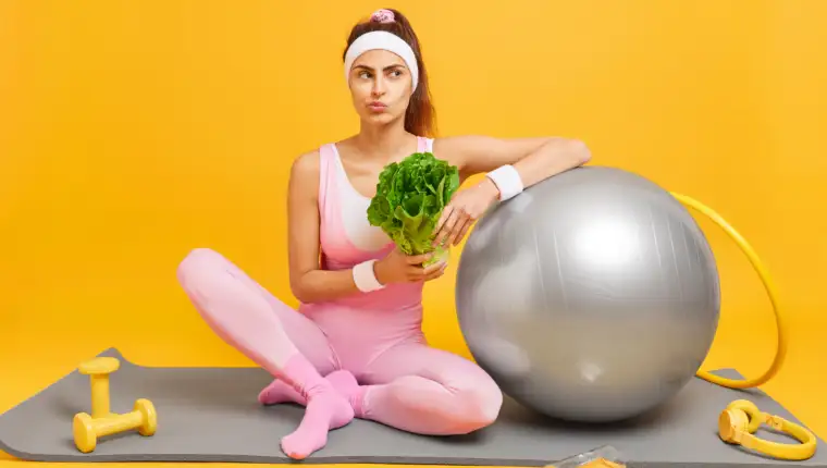 Healthy slim woman keeps to diet has regular fitness training for keeping fit holds green vegetable dressed in sportsclothes sits on mat with headphones dumbbells swiss ball hula hoop in gym