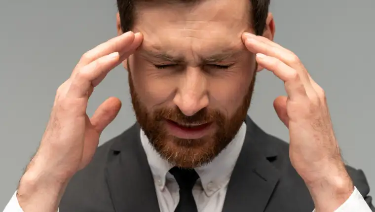 Unhappy frustrated bearded man wearing official style suit massaging temples feeling headache, suffering migraine or high blood pressure. Indoor studio shot isolated on grey background