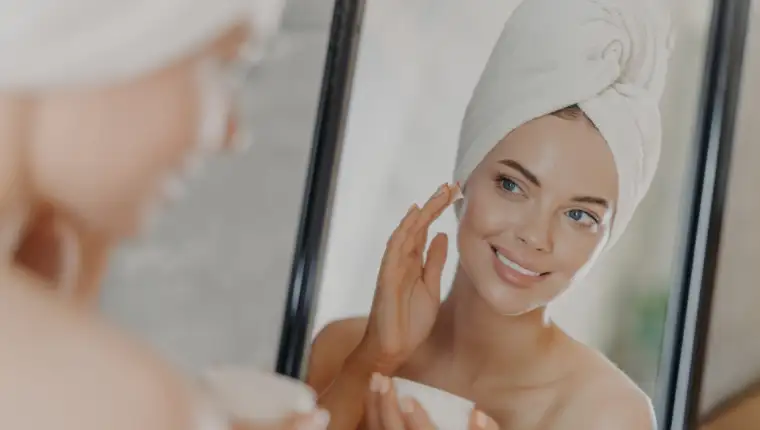 Cute smiling young woman applies beauty cream on face, smiles pleasantly, has minimal makeup, wears wrapped towel on head, looks in mirror, poses in bathroom. Wellness, beauty treatments concept