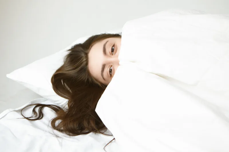 Bedtime, rest and relaxation concept. Playful attractive young woman with beautiful eyes and dark hair relaxing on white pillow, hiding herself behind blanket, looking at camera with happy expression