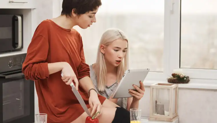 Portrait of two attractive women sitting in kitchen and reading something in tablet, expressing curiosity and interest while preparing salad. Girls passing test on how good they know each other