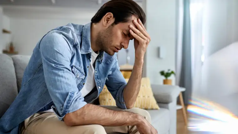 Young man holding his head in pain while sitting on the sofa.