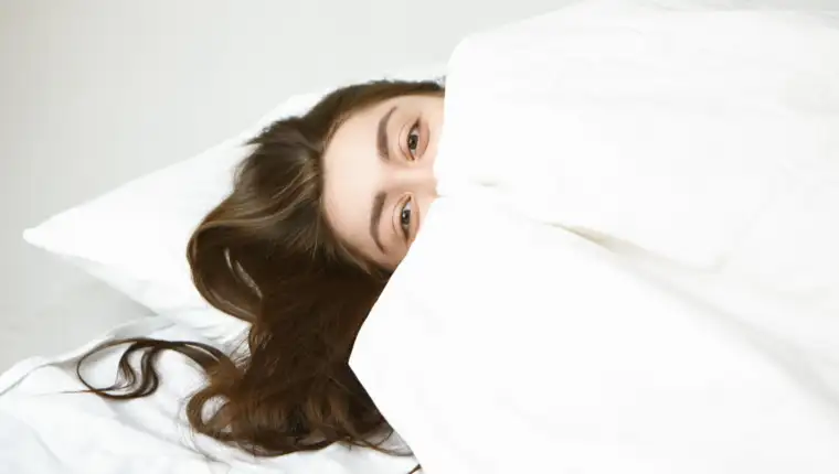 Bedtime, rest and relaxation concept. Playful attractive young woman with beautiful eyes and dark hair relaxing on white pillow, hiding herself behind blanket, looking at camera with happy expression