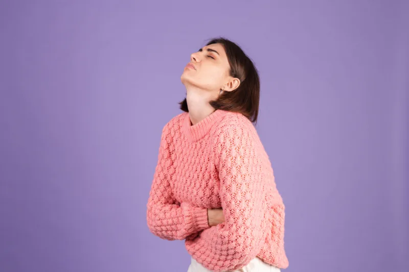Young brunette in pink sweater isolated on purple background suffering stomach ache with painful grimace, feeling sudden period cramps, gynecology concept