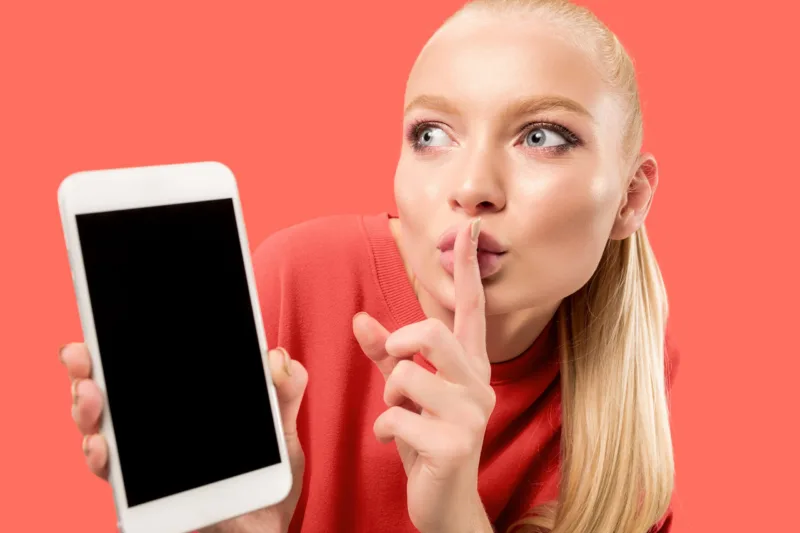 Portrait of a confident casual girl showing blank screen mobile phone isolated over coral background
