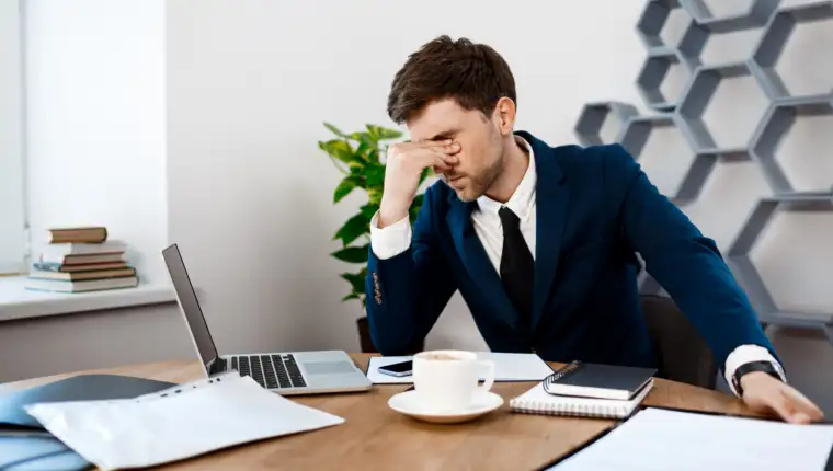 Upset young businessman sitting at workplace, office background.