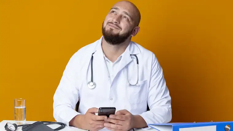 looking at up pleased young bald male doctor wearing medical robe and stethoscope sitting at work desk with medical tools holding phone isolated on orange background