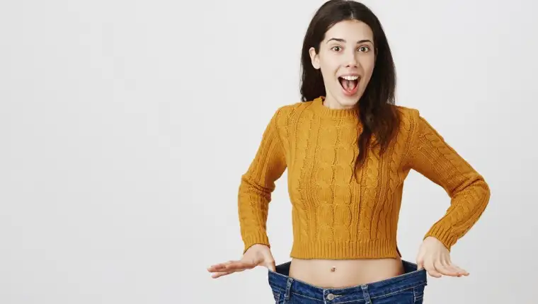 Astonished and happy young lady being impressed and excited because of losing weight, showing empty space in jeans by stretching it, standing over gray background. Girl is ready for summer