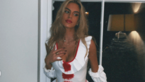 Kćerka Pepa Guardiole Maria Guardiola u bijelom kostimu nalik na medicinske sestre ali sa dubokim dekolteom i crvenim detaljima plava duga kosa desnu ruku drži na grudima lijeva pružena iza nje prozor i zid noćna slika