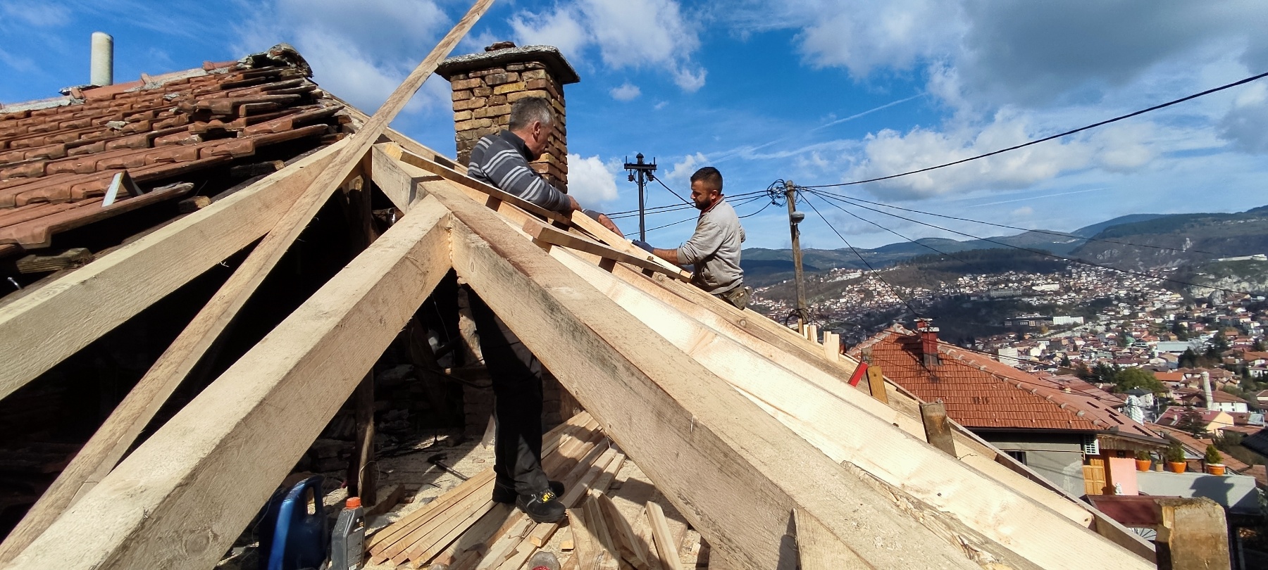 na Širokači je u subotu izgorio krov kuće radnici popravljaju krov stavljaju nove grede u daljini se vidi grad kuće u mahalama plavo nebo dimnjak