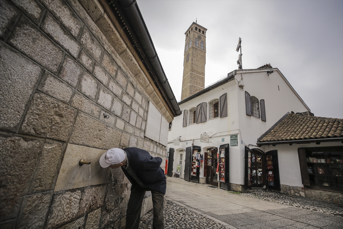 Stare česme simbol Sarajeva česma kod Begove džamije stariji čovjek naget na česmu pije bijeli kačket na glavi ruke na leđima prazna ulica iza njega dan