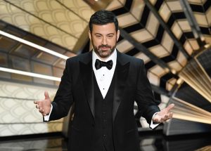 Jimmy Kimmel domaćin 95. dodjele Oscara na sceni iza njega zlatno crni ukrasi Jimmy Kimmel u crnom fraku bijeloj košulji i nosi crnu leptir mašnu ima bradu i brkove tamna kosa ruke dignute napola rašireni dlanovi noć