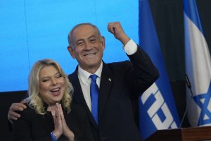 Izraelski opozicioni blok predvođen Netanyahuom osvojio većinu na izborima u Izraelu crni sako bijela košulja plava kravata jednom rukom grli plavu ženu oboje se smiju iza plava pozadina i izraelska zastava