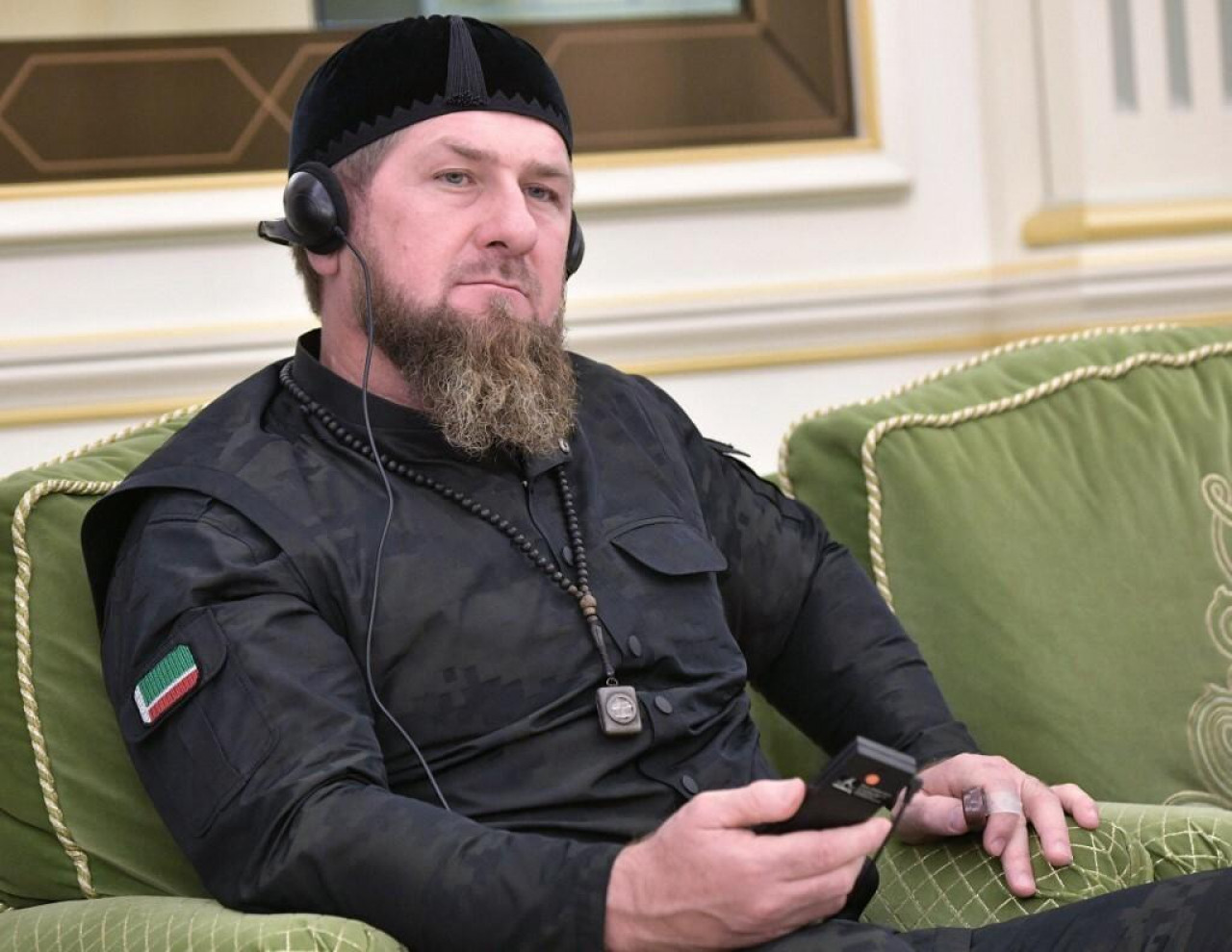 Putinov poslušnik Ramzan Kadirov sjedi u zelenoj fotelji odjeven u crno crna kapa brada na ušima slušalice u ruci aparat za prebacivanje prijevoda iza slika bijeli zidovi