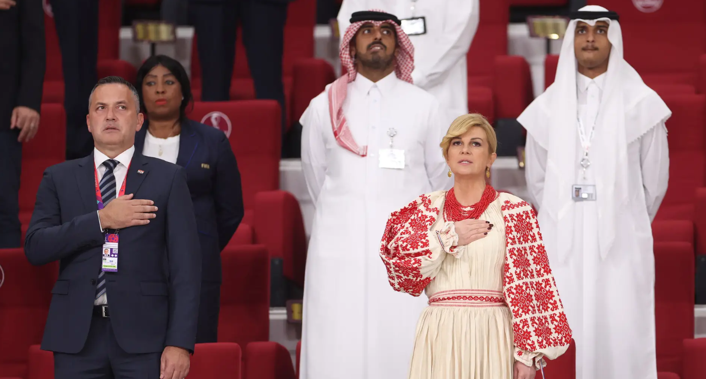 Kolinda Grabar-Kitarović u narodnoj nošnji na tribinama u Dohi gleda utakmicu Hrvatske drži ruku na srcu stoji pored nje muškarac u sivom odijelu tribine crvene stolice iza muškarac u araprskoj nošnji