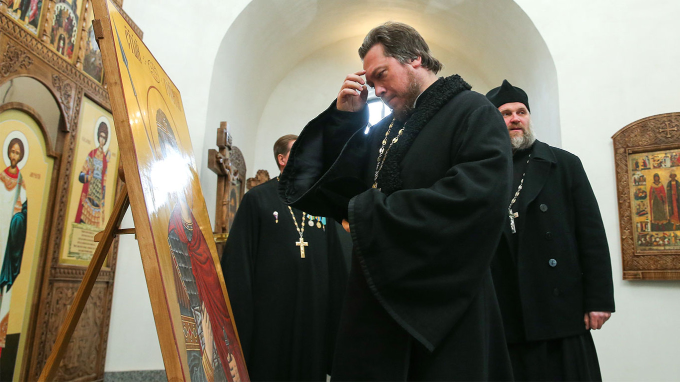 kontroverzni ruski svećenik Mihail Vasiljev stoji ispred ikone i drži prst na čelu sav u crnom lanac sa krstom a vratu iza njega dva svećenika u crnom lanci sa krstom u prostoriji iza se vidi luk na zidu svjetlo upaljeno