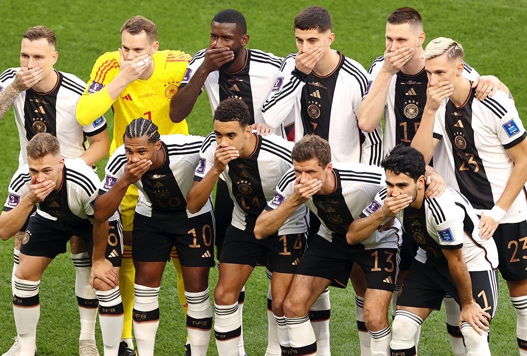 Nijemci prije utakmice iz protesta stavili ruke preko usta fudbaleri Njemačke u bijelo crnim dresovima poziraju i drže ruke preko usta žuti dres na golmanu stoje na travi dan pola tima čiči pola u drugom redu stoji iza njih bijele štucne