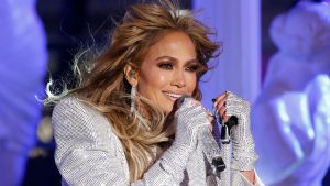 Jennifer Lopez pjeva u haljini na šljokice bijela bijele rukavice sa šljokicama svijetlosmeđa kosa naušnice sa kamenčićima bijeli nokti sa cirkonima našminkana jako plava svjetlost iza nje na bini