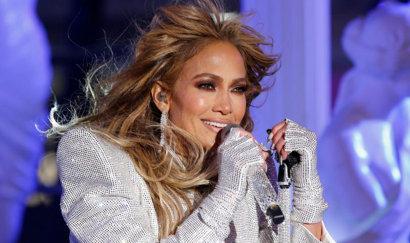 Jennifer Lopez pjeva u haljini na šljokice bijela bijele rukavice sa šljokicama svijetlosmeđa kosa naušnice sa kamenčićima bijeli nokti sa cirkonima našminkana jako plava svjetlost iza nje na bini