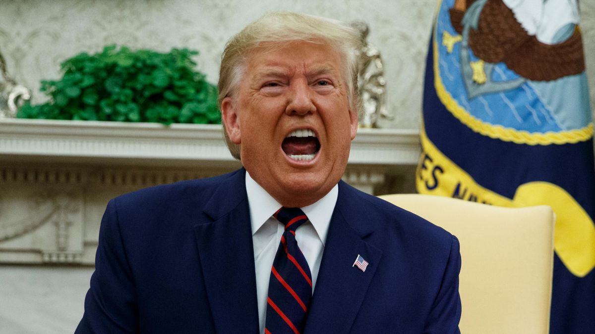 Trump ljut zbog rezultata sjedi u prostoriji plavi sako bijela košulja bordo kravata preplanuo ten usta otvorena kao da viče iza zastava i cvijeće bijeli namještaj